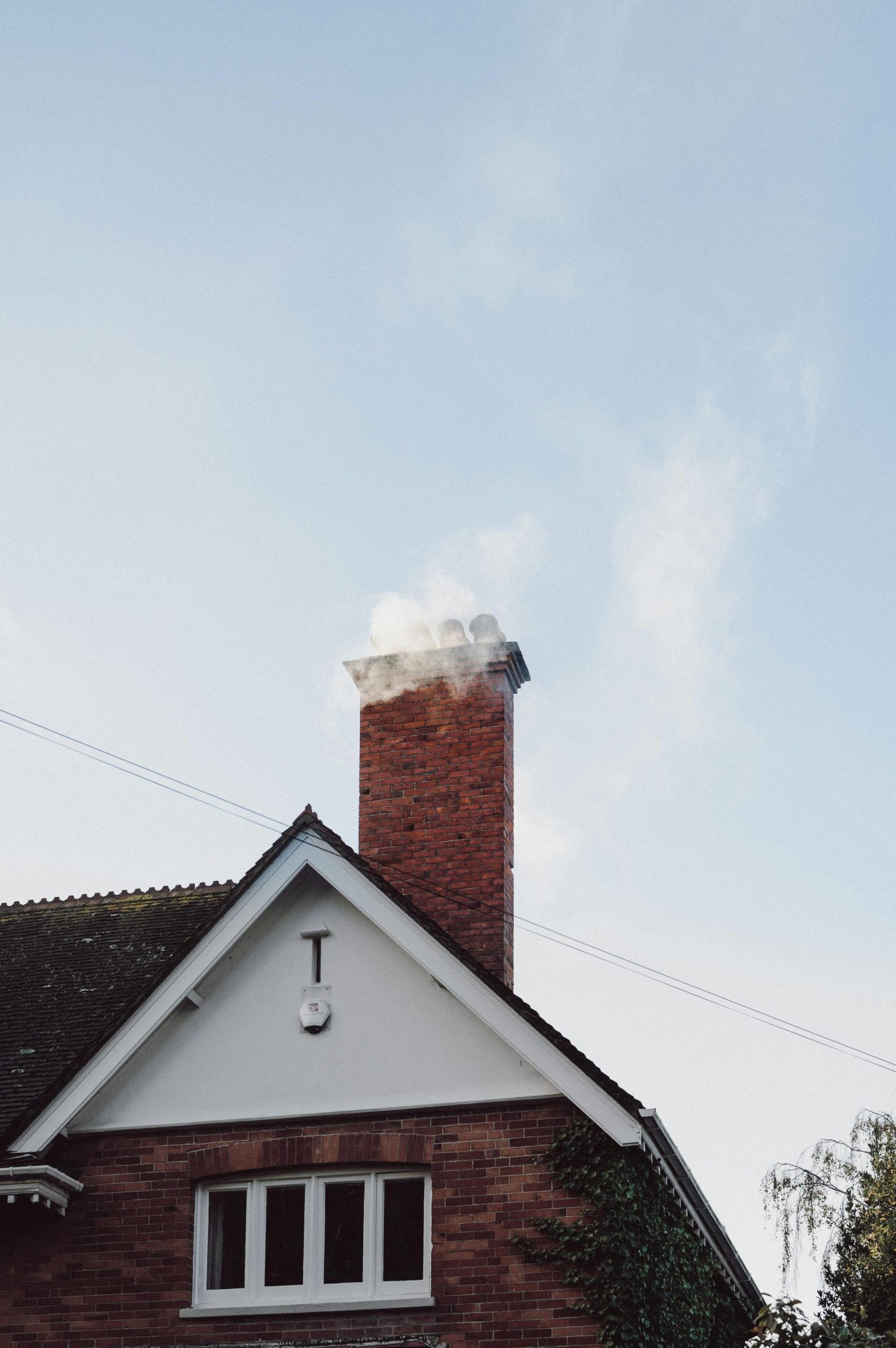 Brick chimney on a house roof with smoke rising into a blue sky.